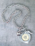 12 gauge shotshell on detailed heart pendant and hung on a toggle clasp with stainless steel paperclip chain. Necklace is displayed on a gray stone background.