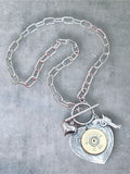 12 gauge shotshell on detailed heart pendant and hung on a toggle clasp with stainless steel paperclip chain. Necklace is displayed on a gray stone background.