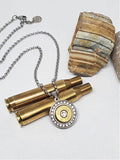 A stainless steel necklace with a round pendant featuring a diamond encrusted solid back and a brass 45 Colt bullet casing in the center, with a diamond crystal in the center of the bullet. The necklace is displayed against two empty brass rifle casings. 