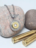 A stainless steel necklace with a round pendant featuring a diamond encrusted solid back and a brass 45 Colt bullet casing in the center, with a diamond crystal in the center of the bullet. The necklace is displayed against a white background with a rolo link chain and a lobster closure.