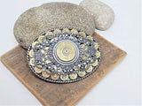 An oval-shaped belt buckle featuring a detailed design with 9mm brass casings and a central 12 gauge shotshell, surrounded by a silver and gold-colored metal filigree on a rope trim.