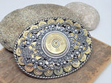 An oval-shaped belt buckle featuring a detailed design with 9mm brass casings and a central 12 gauge shotshell, surrounded by a silver and gold-colored metal filigree on a rope trim.