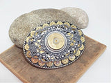An oval-shaped belt buckle featuring a detailed design with 9mm brass casings and a central 12 gauge shotshell, surrounded by a silver and gold-colored metal filigree on a rope trim.