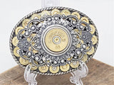An oval-shaped belt buckle featuring a detailed design with 9mm brass casings and a central 12 gauge shotshell, surrounded by a silver and gold-colored metal filigree on a rope trim.