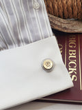 White cuff of a shirt that is gray, black and white striped on the sleeve, showing a brass Winchester 45 Colt bullet casing cufflink.  A burgundy book with "Big Bucks", an antler and some rope are part of the background.