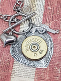 Super close up photo of a heart pendant mounted with a 12 gauge shotshell that has a puffy heart charm and pistol charm flanking it and they are all hung on a smooth toggle clasp.  Pendant is displayed on a red and off white striped vintage looking newsprint.