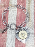 Paperclip style chunky chain bracelet with a spring clasp closure.  Bracelet has heart and pistol charms and a focal heart pendant with a 20 gauge brass shotshell.  Bracelet is displayed on a distressed red and off white newsprint style background.