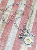 12 gauge shotshell heart necklace with a paperclip style chain and flanked with a small puffy heart charm and pistol charm.  Necklace is displayed on a red and off white striped vintage looking newsprint.