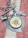 Super close up photo of a heart pendant mounted with a 12 gauge shotshell that has a puffy heart charm and pistol charm flanking it and they are all hung on a smooth toggle clasp.  Pendant is displayed on a red and off white striped vintage looking newsprint.