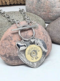 Close up of a 12 gauge Winchester brand shotshell mounted to a detailed heart pendant with a puffy heart and pistol charm all hanging from a stainless smooth toggle clasp.  Necklace is displayed against a pink stone with other stones in the background.