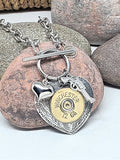 Close up of a 12 gauge Winchester brand shotshell mounted to a detailed heart pendant with a puffy heart and pistol charm all hanging from a stainless smooth toggle clasp.  Necklace is displayed against a pink stone with other stones in the background.