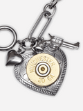 Close up a silver etched heart pendant embellished with a brass Winchester brand 20g shotshell.  It is flanked by smaller pistol and puffy heart charms.  Laying on a white background.