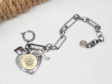 Flay lay of a paperclip style chain bracelet that has a heart charm mounted with a brass 20 gauge Winchester brand shotshell head and flanked by two smaller charms - a pistol and a puffy heart.  Bracelet has a large spring clasp.  Background is white with rope showing in the background.