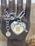 Paper clip chain style bracelet with heart and pistol charms and a focal heart pendant with a 20 gauge brass shotshell.  Bracelet is displayed on a metal hand display to show the details up close. 