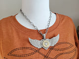 Large winged heart necklace with pavé crystal pendant, brass 12 gauge shotshell center, and chunky silver paperclip chain shown on mannequin wearing a dark orange t-shirt with black boot stitch design on front.