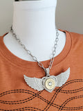 Large winged heart necklace with pavé crystal pendant, brass 12 gauge shotshell center, and chunky silver paperclip chain shown on mannequin wearing a rusty orange t-shirt with black boot stitch detailing.