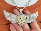 Close-up view of a pave encrusted diamond winged heart necklace with a brass shotshell as the focal piece and a large diamond crystal in center.  Pendant is being held in a hand while it is being worn by a mannequin wearing an orange t-shirt.