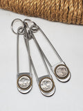 A group of three stainless steel jumbo safety pins mounted with genuine vintage buffalo nickel coins that are mounted in antique brass rope edge bezels. Displayed on a white background with a roll of rope in background.