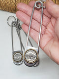 Hand holding a jumbo safety pin that has a genuine buffalo nickel coin mounted in a brass rope edge bezel for size and scale.