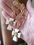 A pair of gold and white howlite cross toggle necklaces with bullet casings in center held in a person's hand.