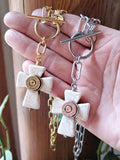Two white howlite cross necklaces held in a hand. One features gold paperclip chain and toggle clasp, and the other a silver paperclip chain and toggle clasp, both with matching bullet casings in the center.