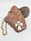 A handmade howlite cross pendant necklace with gold over stainless steel paperclip chain, featuring a white howlite cross and a brass bullet casing mounted in the center, displayed on a pinkish-brown stone against a white background.
