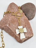 A close up photo of a handmade howlite cross pendant necklace with gold over stainless steel paperclip chain, featuring a white howlite cross and a brass bullet casing mounted in the center, displayed on a pinkish-brown stone against a white background.