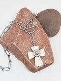 A close up photo of a handmade howlite cross pendant necklace with stainless steel paperclip chain, featuring a white howlite cross and a nickel silver bullet casing mounted in the center, displayed on a pinkish-brown stone against a white background.
