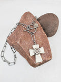 A white howlite cross pendant necklace with stainless steel paperclip chain, featuring a nickel 45 auto bullet casing mounted in the center, displayed on a pinkish-brown stone against a white background.