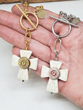 Two white howlite cross necklaces with bullet casings mounted in the center.  Suspended from smooth toggle clasps and displayed over a hand.