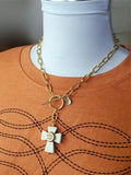 Gold over stainless steel white howlite cross necklace with a brass bullet casing mounted in the center and suspended from a large round toggle clasp also in gold over stainless steel.  Displayed on a mannequin wearing a boot stitch detailed rust or orange t shirt.