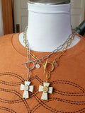 Gold over stainless steel and a stainless steel white howlite cross necklaces with brass and nickel bullet casings respectively mounted in the centers of those crosses and suspended from large round toggle clasps in the same respective metal colors.  Displayed on a mannequin wearing a boot stitch detailed rust or orange t shirt.