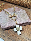 A white howlite cross paperclip chain with toggle clasp gold over stainless steel necklace.  The howlite cross has a brass bullet casing in the center.  Necklace is displayed over a pinkish mauve flat stone sitting on a wood base with rope in the background.