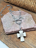 A white howlite cross paperclip chain with toggle clasp stainless steel necklace.  The howlite cross has a nickel bullet casing in the center.  Necklace is displayed over a pinkish mauve flat stone sitting on a wood base with rope in the background.