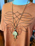 Long brass fish pendant necklace worn on mannequin with chunky gold paperclip chain and front ring fastener.  Mannequin is wearing an orange or rust colored t-shirt that has black boot stitching motif across front.