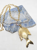 Large solid brass fish pendant necklace displayed on blue stone with gold paperclip chain and vintage ring fastener.