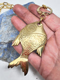 Close-up of solid brass fish pendant held in hand showing stamped scale detail, fins, eye, and warm brass finish.