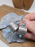 Pair of authentic aluminum goose band earrings held in hand to show scale, curved band construction, turquoise beads and lightweight dangle design.