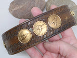 One-of-a-kind Key of A brown leather cuff bracelet held in hand, showing three round vintage bronze Ducks Unlimited Sponsor pins featuring Snow Goose, Gadwall and American Widgeon. The repurposed western tooled leather belt has rich natural wear and patina with matching gold-tone hardware.