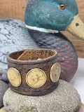 Upcycled Ducks Unlimited Sponsor pin cuff bracelet displayed on a smooth river stone with vintage painted duck decoy in the background. Dark brown western tooled leather contrasts with three warm bronze medallions depicting different waterfowl species.