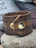 Back view of the upcycled brown leather cuff bracelet showing the overlapping vintage tooled belt ends and shiny gold-tone snap closure. The supple worn leather displays natural aging, softened edges and tooling from its original life as a men's western belt.