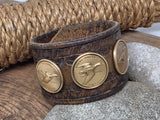 Detailed close-up of the vintage leather cuff resting on stones and weathered wood with a coil of rope in the background, showing the Snow Goose and Gadwall bronze Ducks Unlimited Sponsor medallions and part of the American Widgeon design against richly worn western tooled leather.