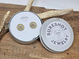 Photo of a silver metal tin containing a pair of Winchester brand brass 28 gauge shotshell studs.  Lid of tin has the SureShot Jewelry logo label.  Tin is leaning on a deer antler and everything is displayed on a wood board.