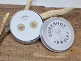 Photo of a silver metal tin containing a pair of vintage Remington-Peters brand brass 28 gauge shotshell studs.  Lid of tin has the SureShot Jewelry logo label.  Tin is leaning on a deer antler and everything is displayed on a wood board.