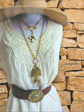 Long brass fish pendant necklace styled on mannequin with other SureShot Jewelry pieces for outdoor-inspired layered jewelry look.  Mannequin is wearing a cream with lace overlay western dress with a tan hat and is standing in front of a tan colored stone wall.